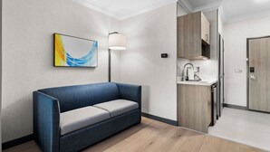 Ein modernes Hotelzimmer mit blauem Sofa, einem Gemälde, einer Stehlampe, einem Holzschrank und einem integrierten Waschbecken.