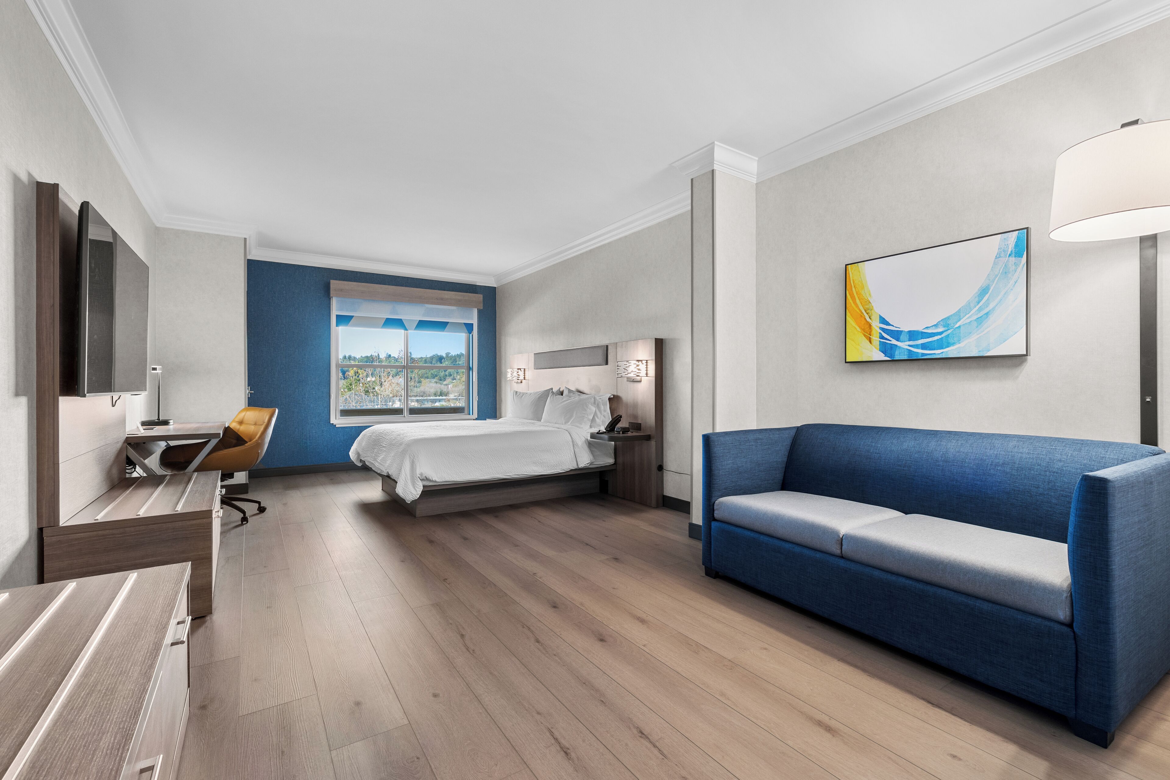 A modern hotel room with a bed, a blue sofa, a desk, and a window with a city view.