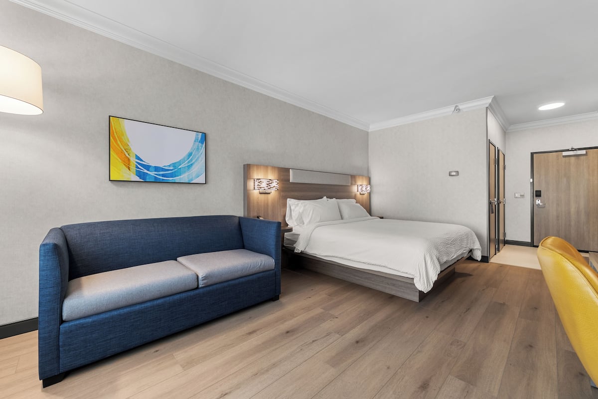 Una habitación de hotel moderna con un sofá azul, una cama con ropa blanca, piso de madera y una puerta de madera.