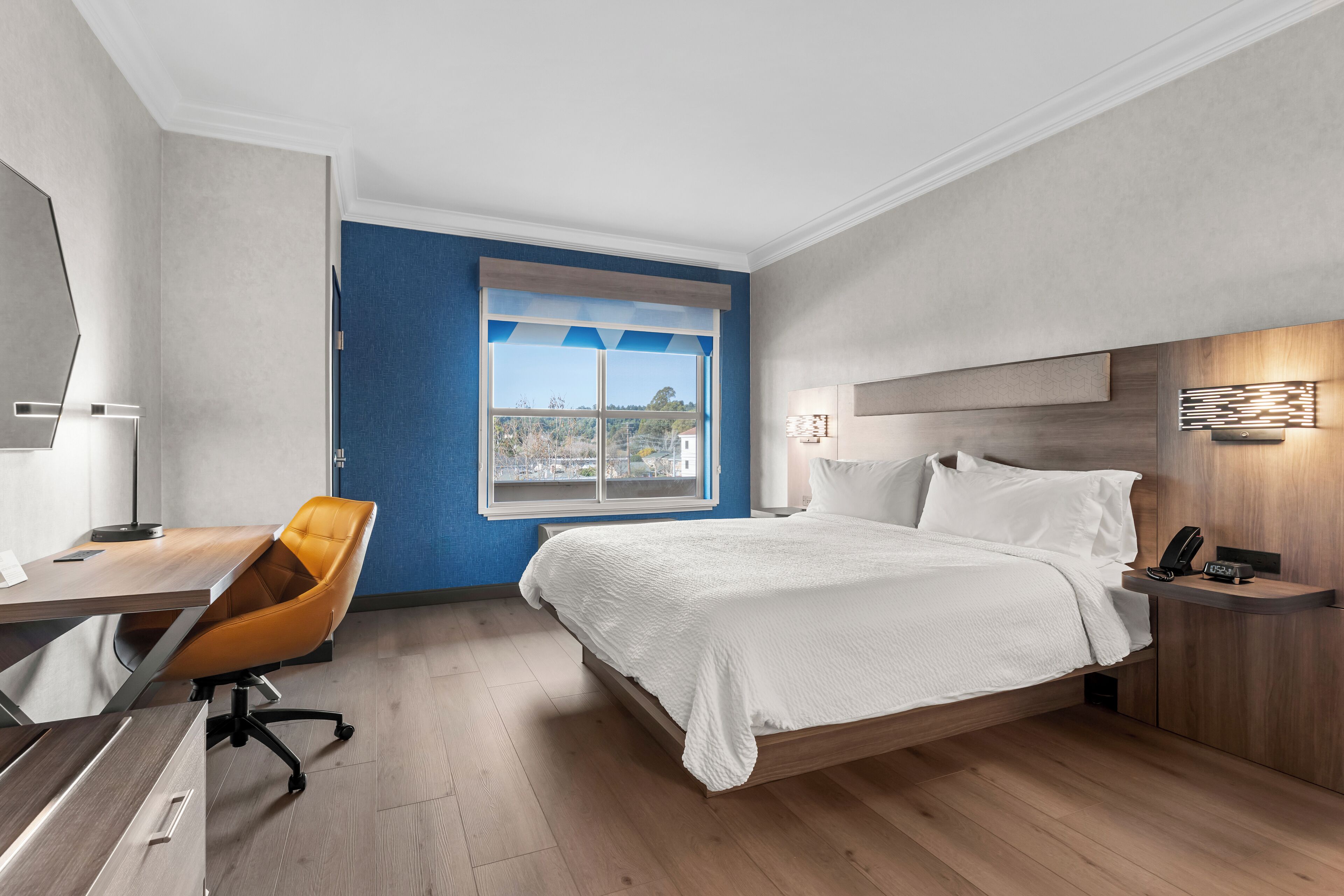 A modern hotel room with a bed, a desk, an orange chair, a nightstand, and a window with a view.