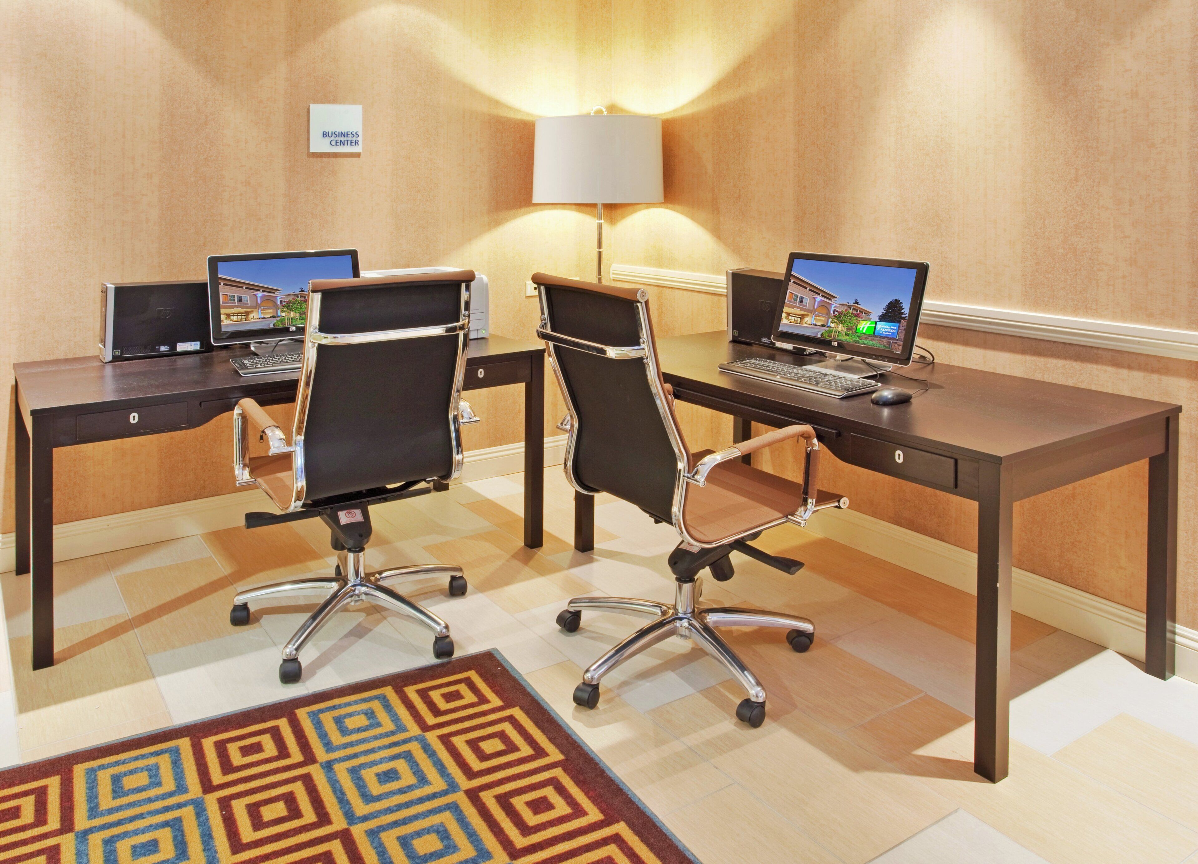 A conference room with two desks, each with a laptop and a chair. There is a lamp on the wall and a carpet with a geometric pattern.