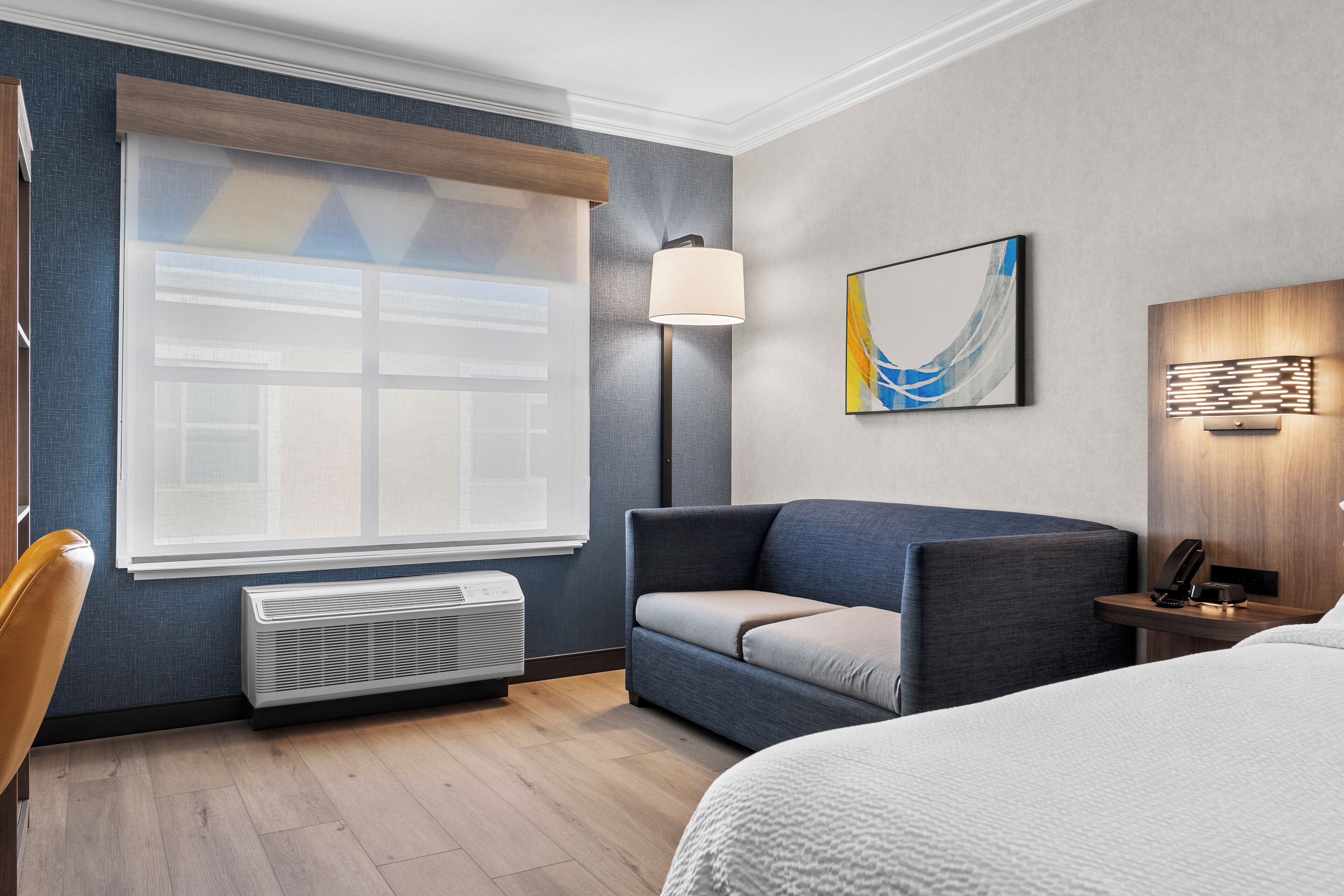 A hotel room with a large window, a sofa, a bed, a bedside table with a lamp, and a wall-mounted artwork.
