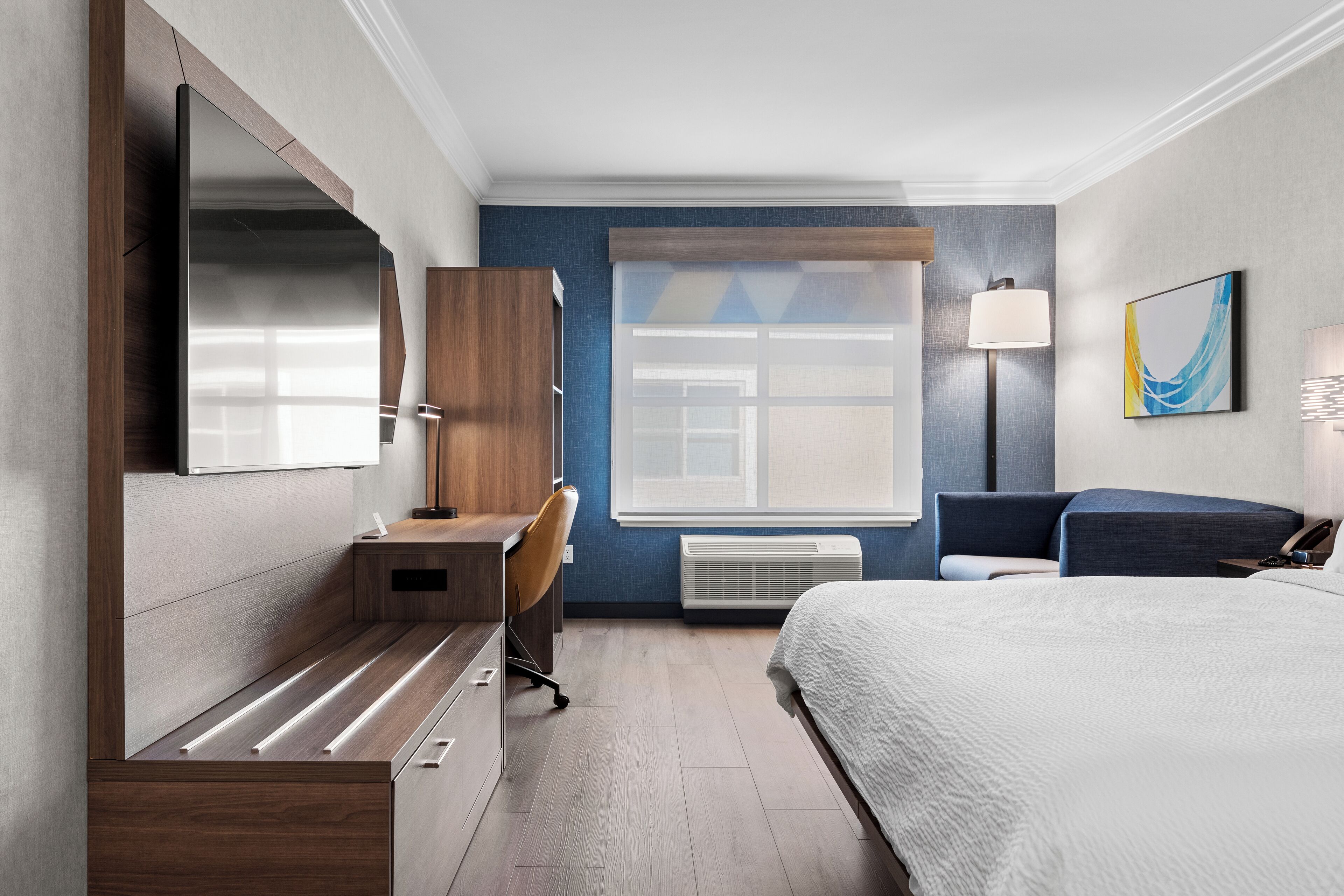 A modern hotel room with a large bed, a desk with a chair, a wardrobe, a large window, and a wall-mounted lamp.