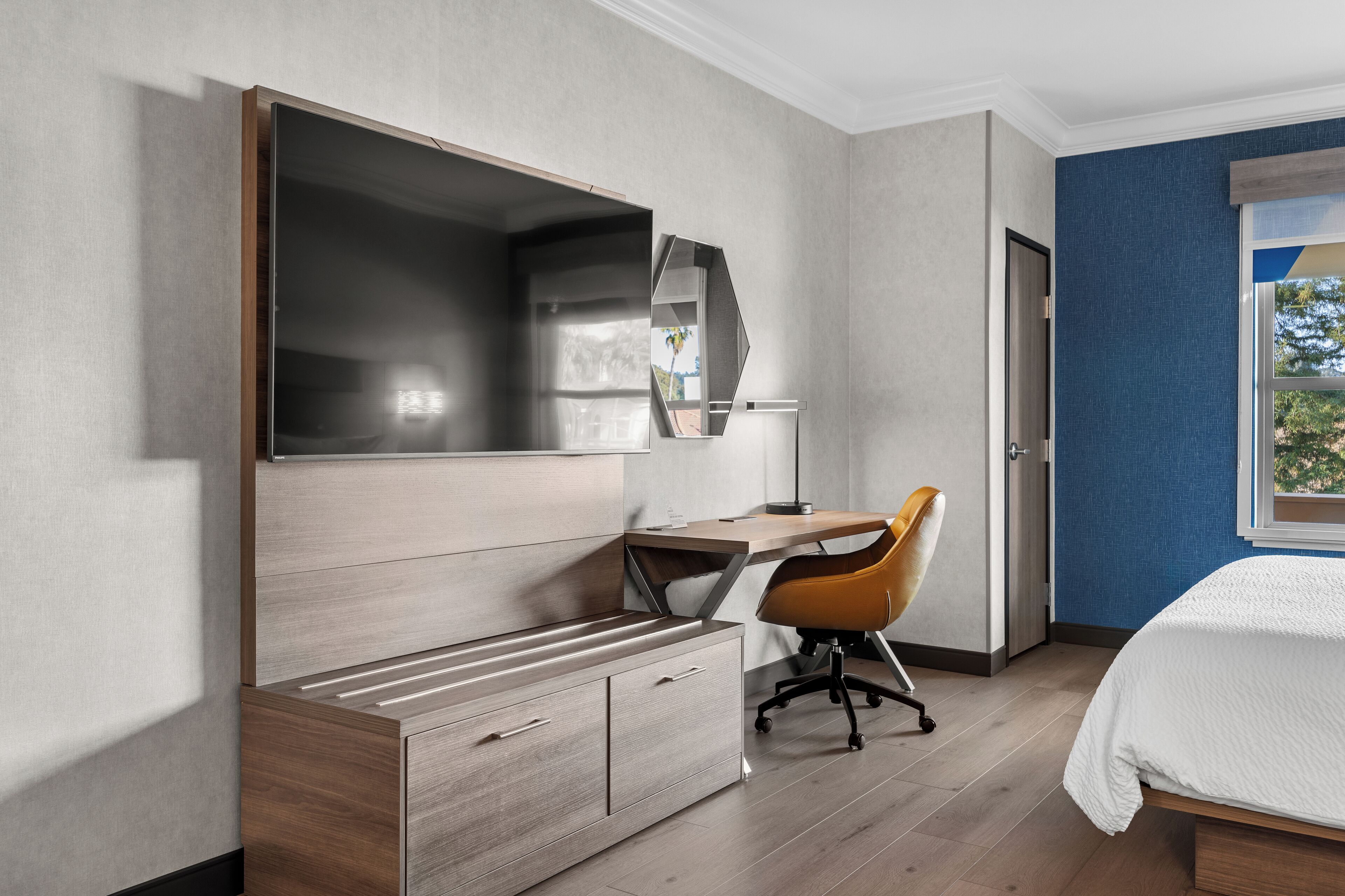 A modern hotel room with a large bed, a desk with a chair, a wardrobe, and a large mirror.