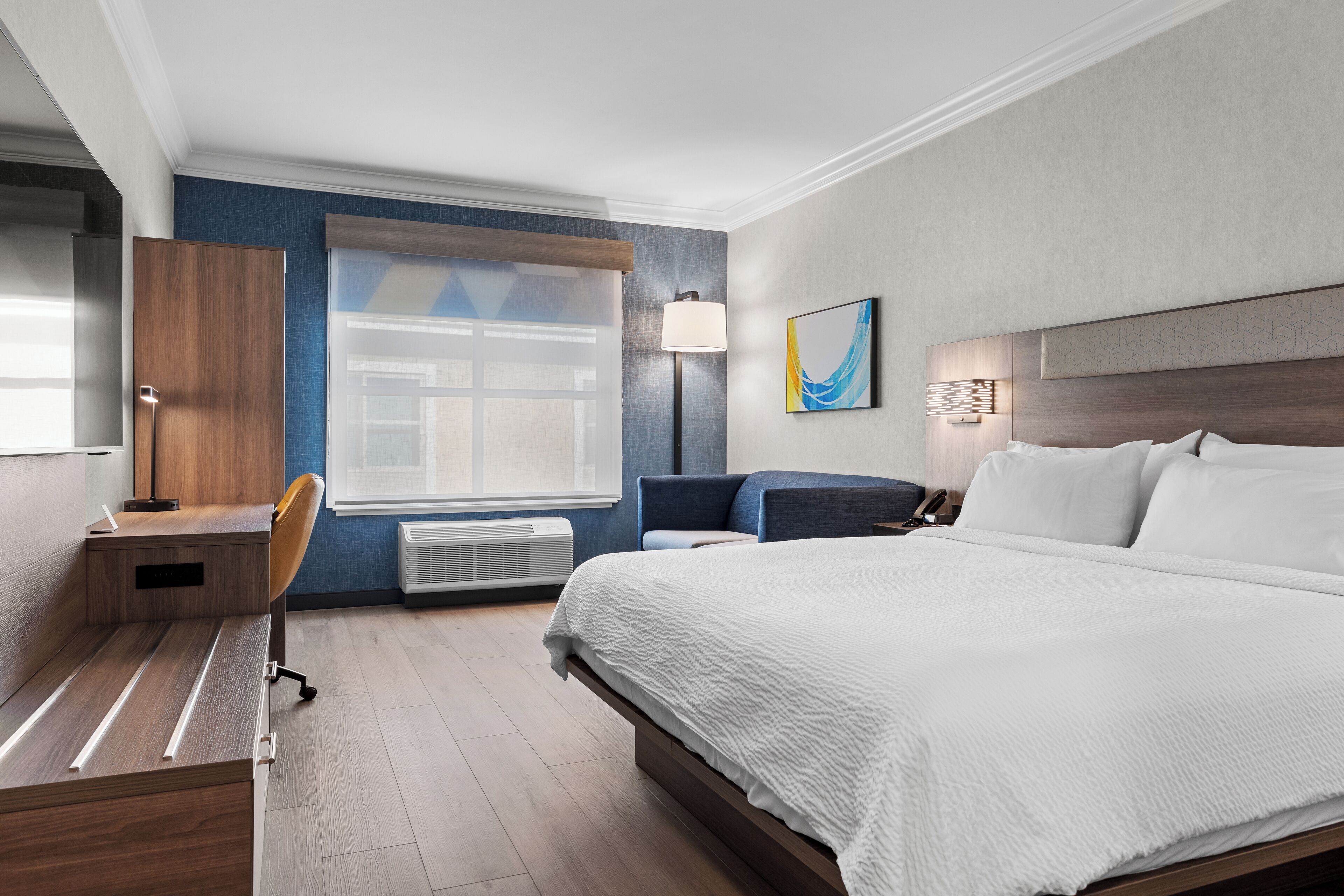 Quarto de hotel moderno com uma cama grande, uma escrivaninha, uma cadeira, um candeeiro de pé e uma janela com persianas.