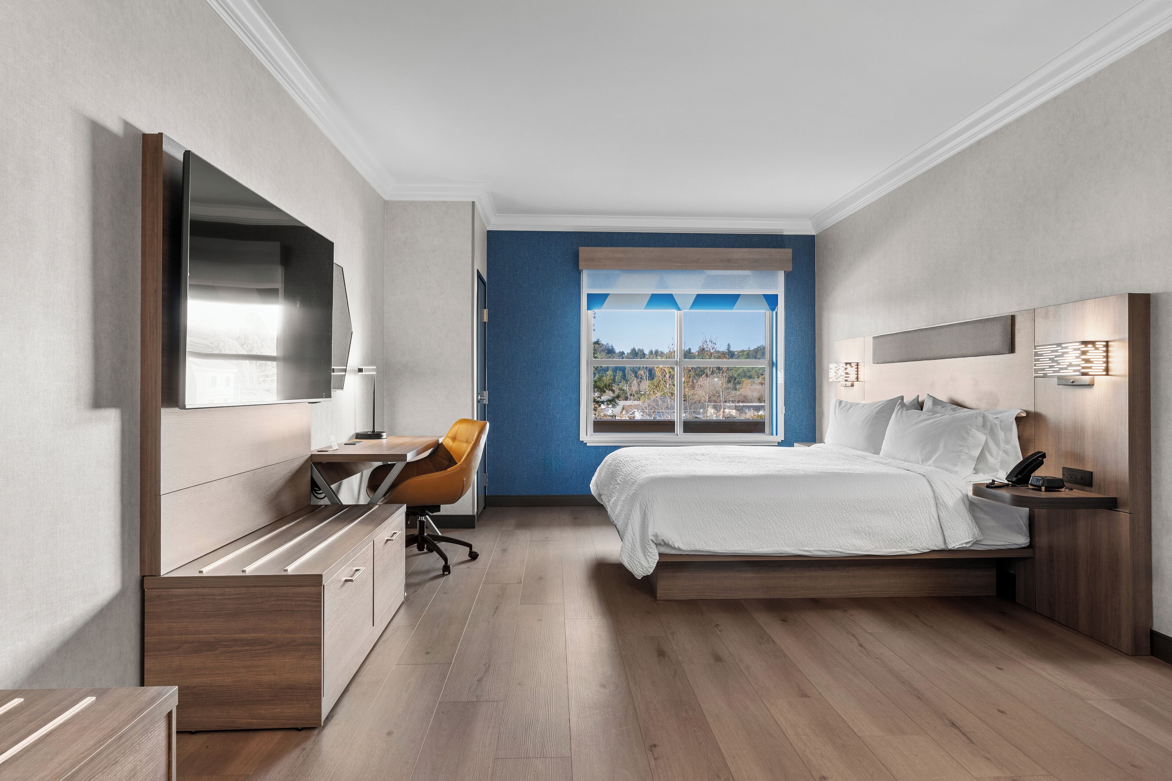 Quarto de hotel moderno com cama, escrivaninha, cadeira e uma televisão grande.