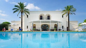 A poolside view of a hotel with a covered outdoor seating area, palm trees, and a clear blue sky. - Masseria la Baronessa Resort (Tuglie)
