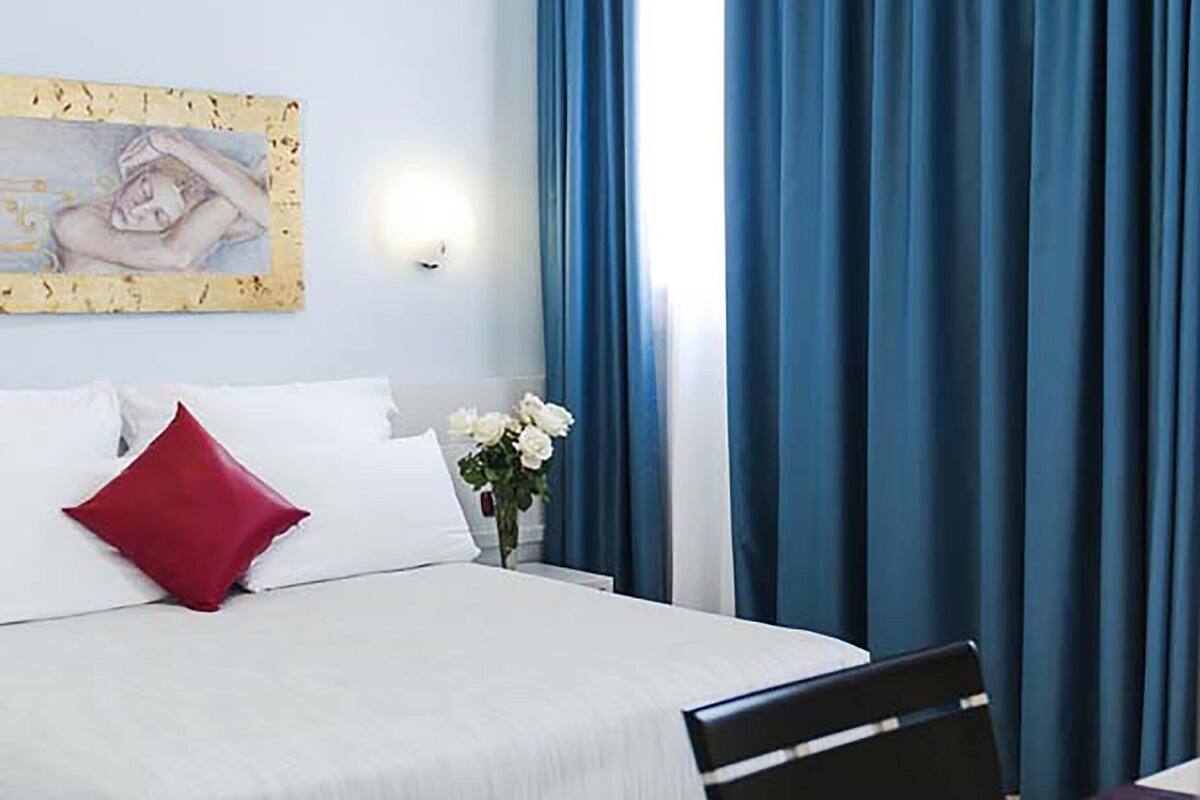 A hotel room with a bed, a desk, a vase with flowers, and blue curtains.