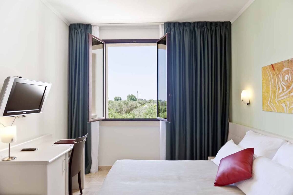A hotel room with a bed, a desk with a computer, a window with curtains, and a painting on the wall.