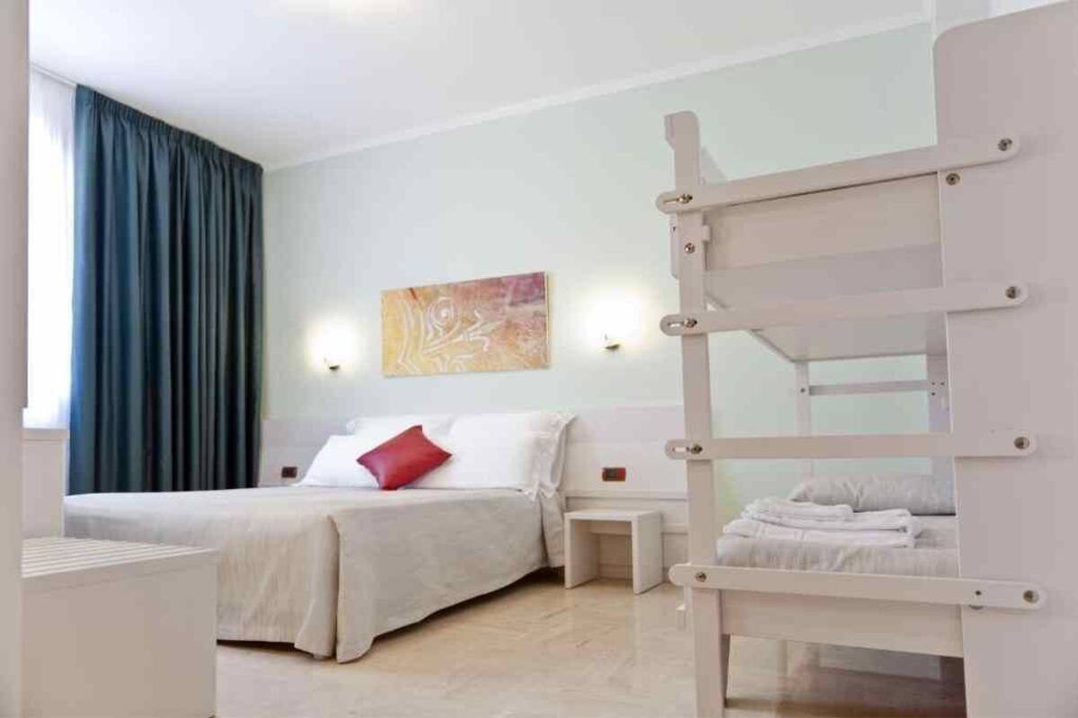 A hotel room with a bunk bed, a bedside table, a wall-mounted light, and a painting on the wall.