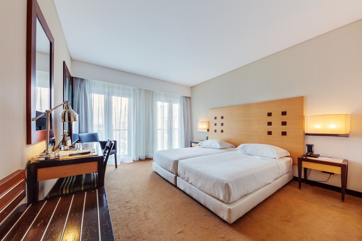 A hotel room with a large bed, a desk with a lamp, a chair, and a bedside table.
