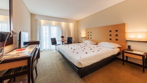 Ein Hotelzimmer mit einem großen Bett, einem Schreibtisch mit Computer, einem Sessel, einem kleinen Tisch mit Vase und einem Fenster mit Vorhängen.