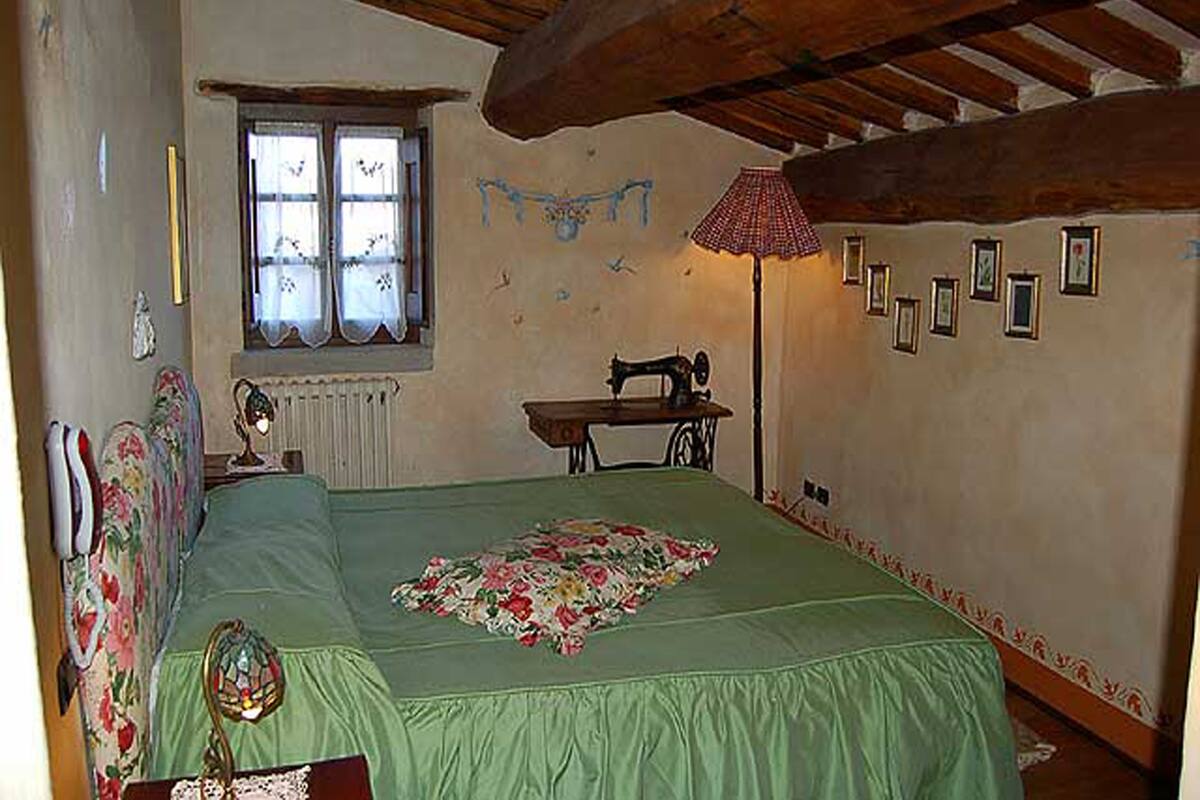 Ein Schlafzimmer mit grüner Bettdecke, einer alten Nähmaschine, einem Fenster mit Spitzenvorhängen und einer Holzbalkendecke.