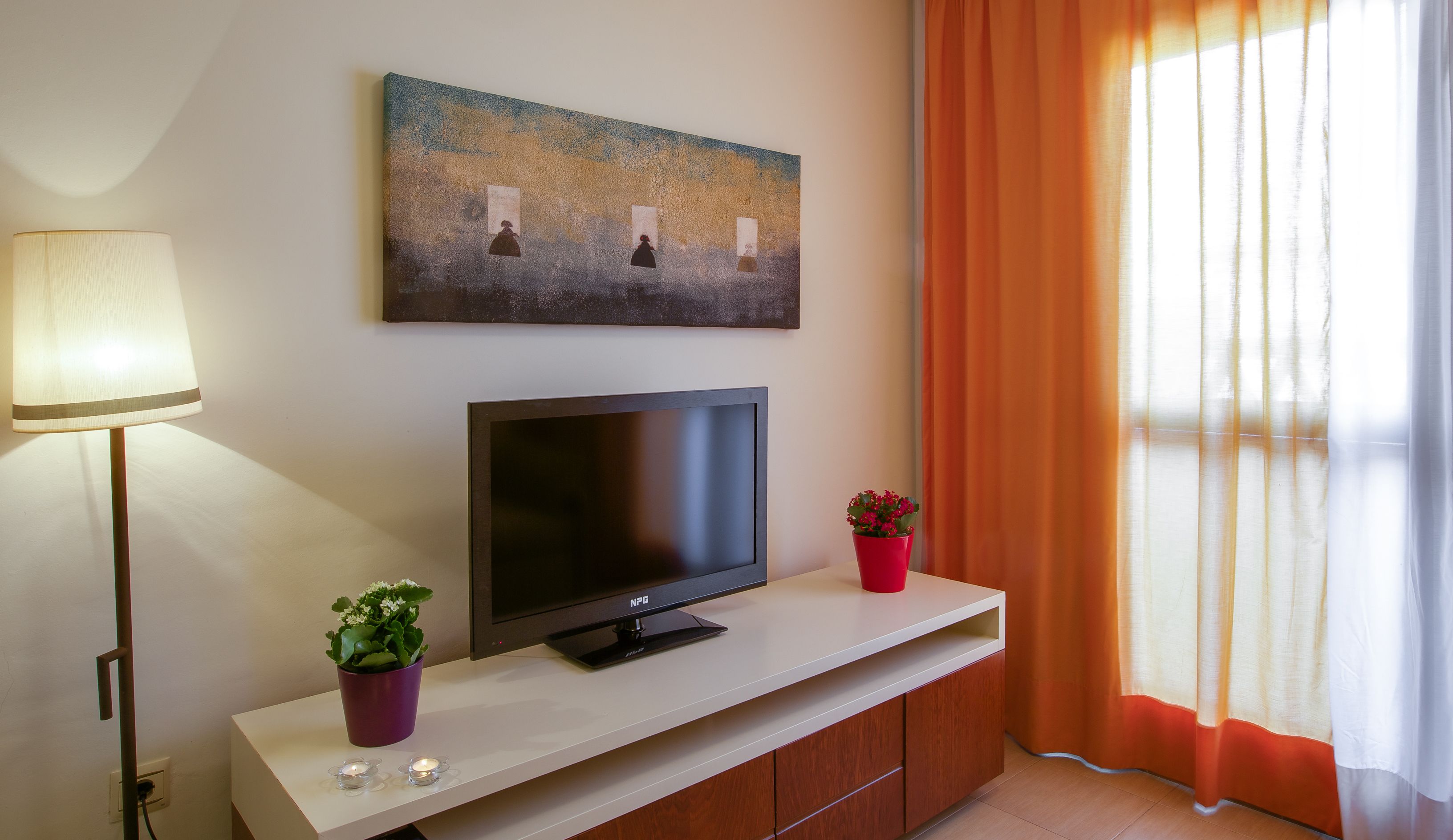 Une chambre d’hôtel moderne avec un téléviseur à écran plat posé sur une console blanche, un tableau au-dessus, une lampe sur la gauche et une fenêtre avec des rideaux orange.
