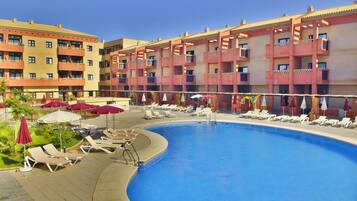 Zona de piscina de hotel con tumbonas y sombrillas, rodeada de edificios de varios pisos con balcones.
