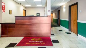 Zona de recepción de un hotel con mostrador, alfombra roja con el nombre del hotel y un pasillo con puertas y una pared verde.
