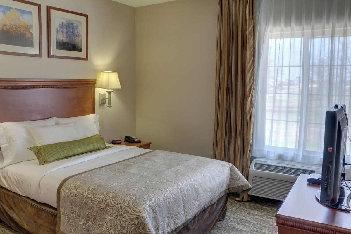 Une chambre d’hôtel avec un lit, un bureau avec un ordinateur, une lampe et une fenêtre avec des rideaux.