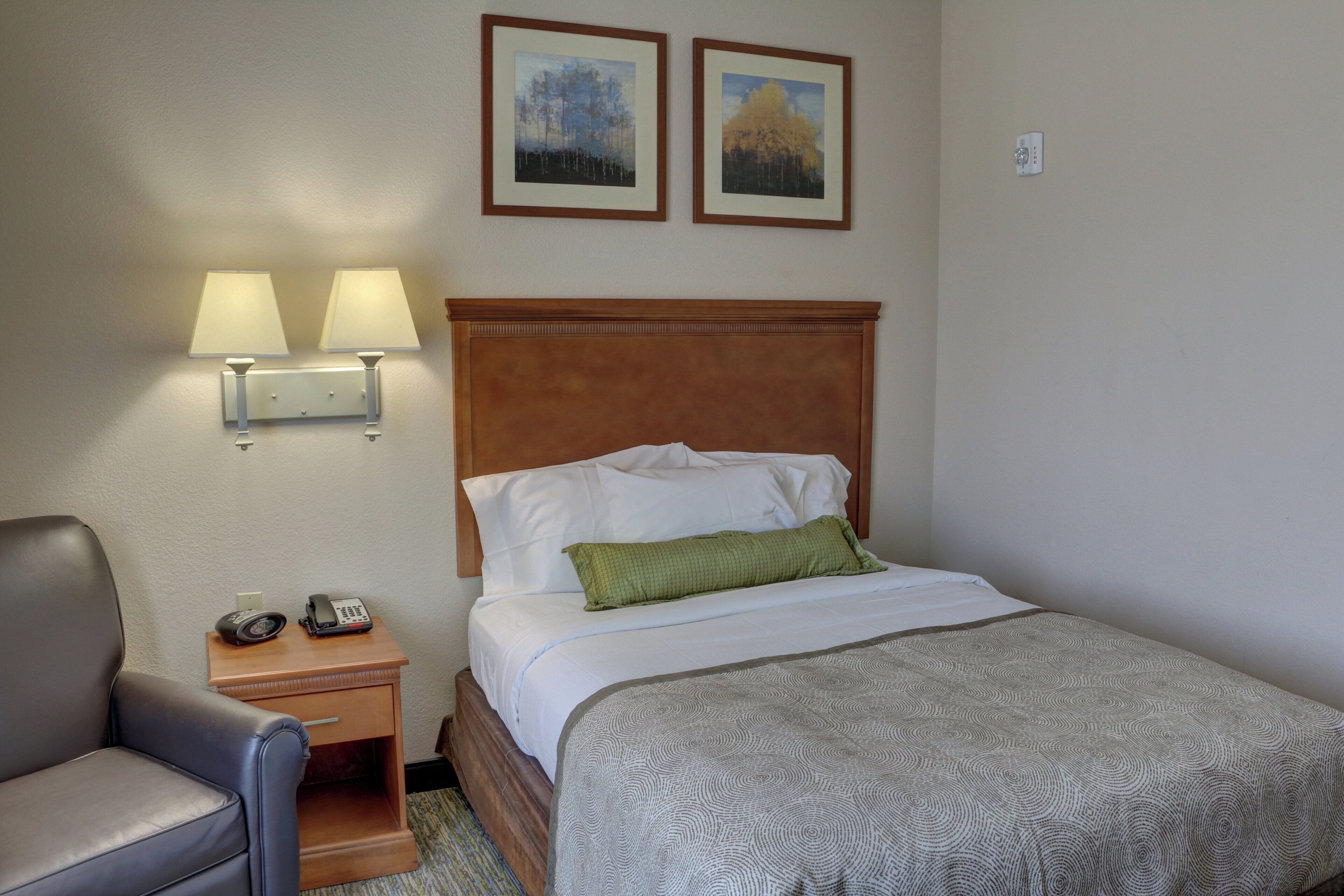 A hotel room with a bed, a chair, a nightstand with a phone, two wall-mounted lamps, and two framed pictures on the wall.