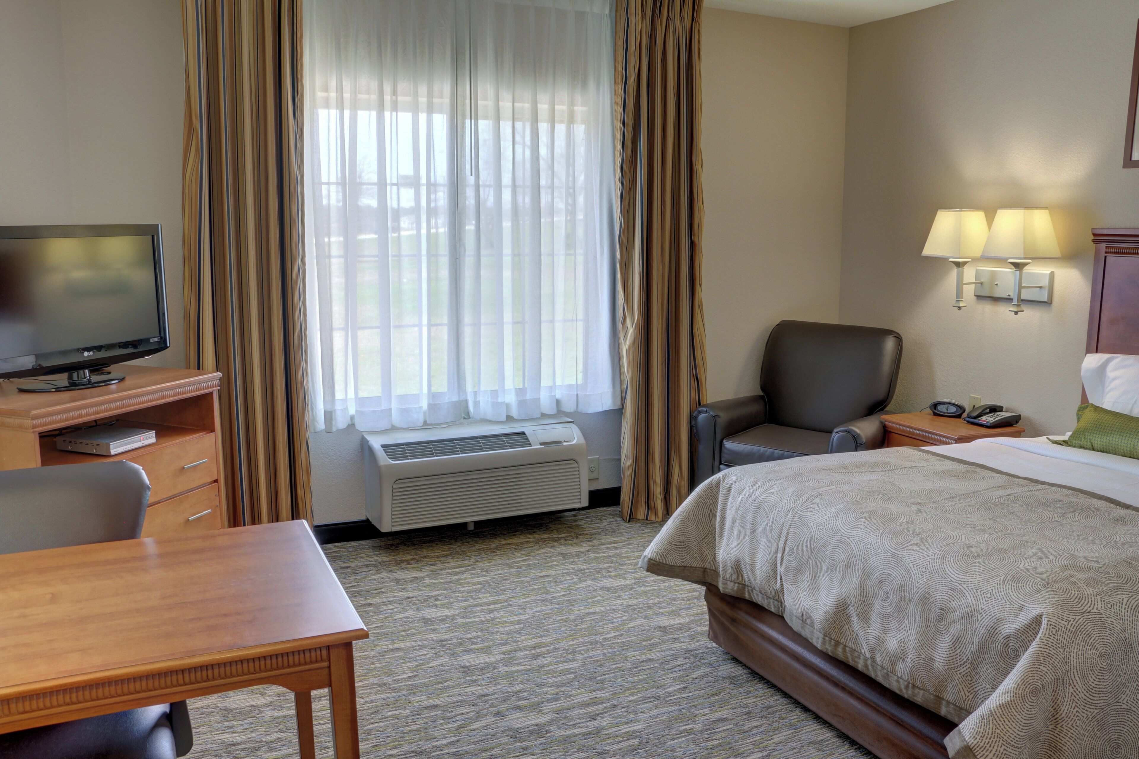 A hotel room with a bed, a desk with a television, a chair, an air conditioning unit, and a window with curtains.