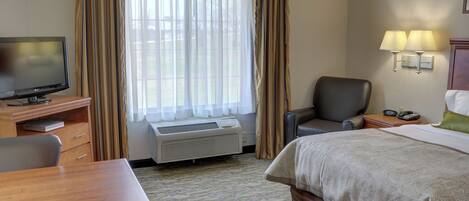 A hotel room with a bed, a desk with a television, a chair, an air conditioning unit, and a window with curtains.
