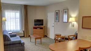 A hotel room with a sofa, a TV, a dining table with chairs, a lamp, and a window with curtains.