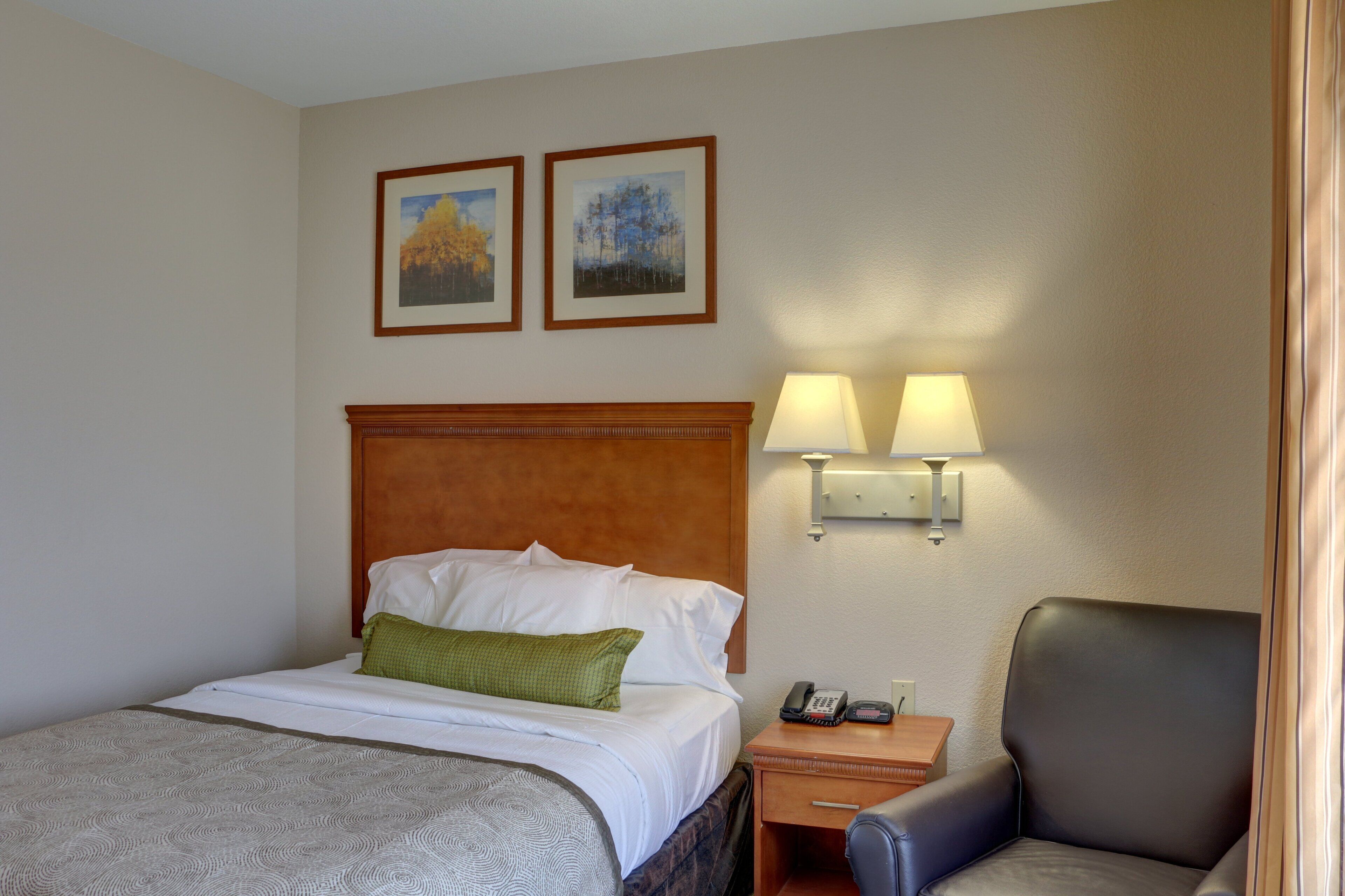 A hotel room with a bed, a nightstand with a lamp, a chair, and two framed pictures on the wall.
