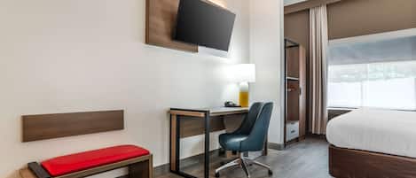 Une chambre d’hôtel comprenant un lit, un bureau avec une chaise, un banc, un téléviseur fixé au mur et une fenêtre avec des rideaux.