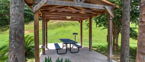 A wooden gazebo with a picnic table, surrounded by trees and grass.