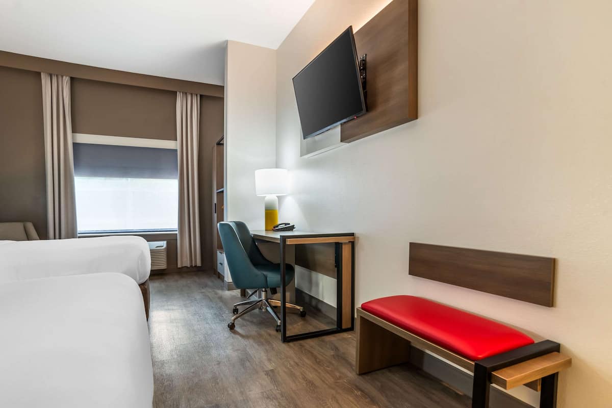 A hotel room with two beds, a desk with a chair, a TV mounted on the wall, and a window with curtains.