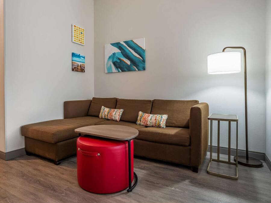 A modern living room with a brown sectional sofa, a red ottoman, a floor lamp, and abstract wall art.