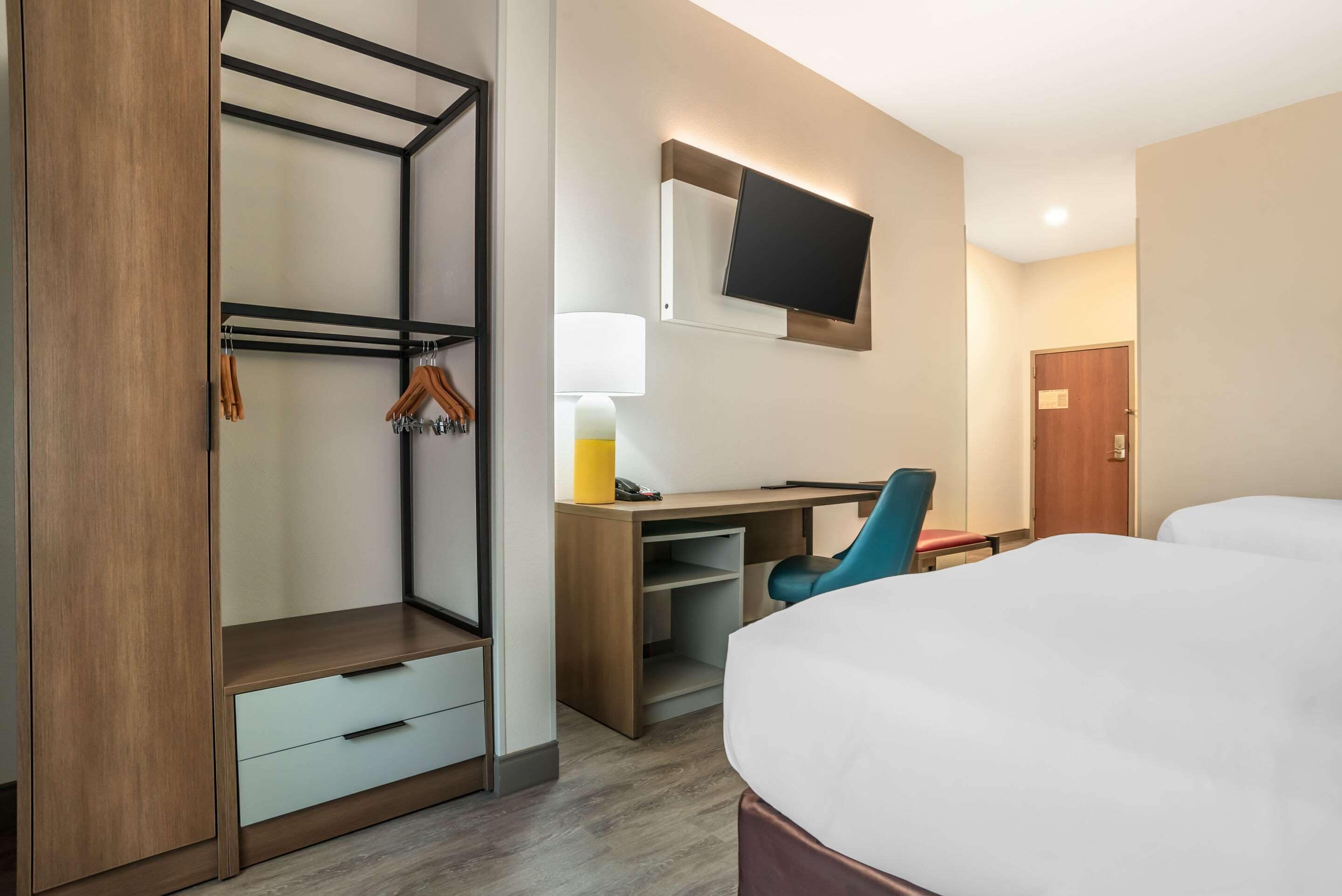 Una camera d'albergo con un letto, una scrivania, una sedia, una TV a parete, un armadio e una porta.