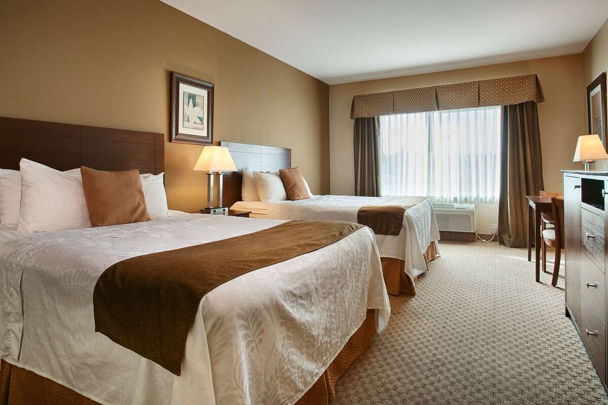 A hotel room with two beds, a desk, a dresser, a TV, and a window with curtains.