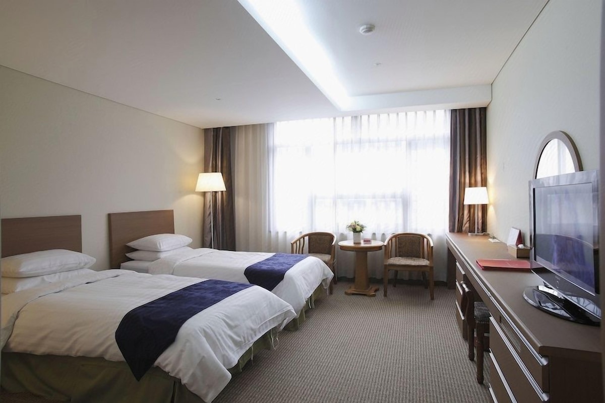 A hotel room with two beds, a desk with a television, a chair, and a small table with a lamp.