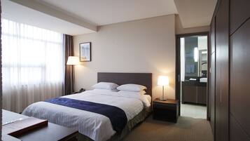 A hotel room with a bed, bedside tables, a desk, a lamp, a window with curtains, and a door leading to a bathroom.