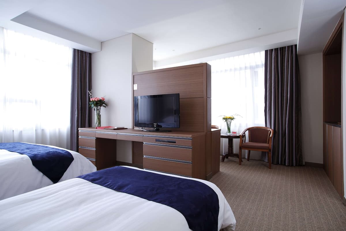 Une chambre d’hôtel avec deux lits, un bureau avec une télévision, une chaise et une petite table sur laquelle est posé un vase de fleurs.