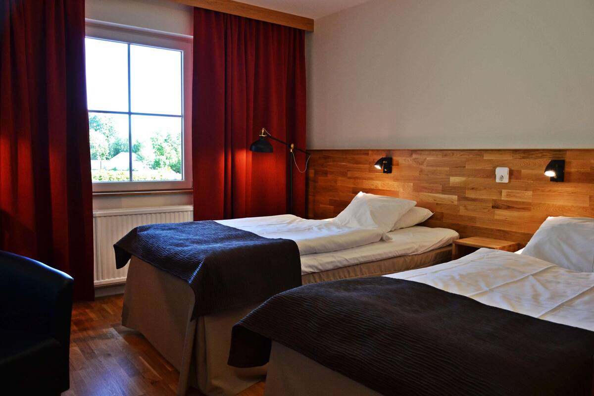 Ein Hotelzimmer mit zwei Betten, einem Fenster mit Vorhängen, einem hölzernen Kopfteil und einem Nachttisch mit Lampe.