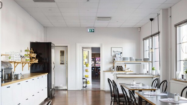 A spacious dining area with wooden tables and chairs, a kitchen area with white cabinets and a black refrigerator, and large windows providing natural light. - Dalhall Hotell (Åmål)
