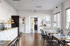 A spacious dining area with wooden tables and chairs, a kitchen area with white cabinets and a black refrigerator, and large windows providing natural light. - Dalhall Hotell (Åmål)