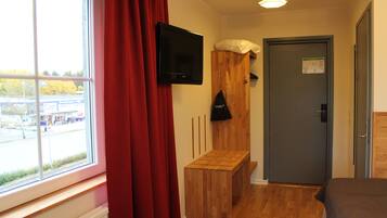 Ett hotellrum med en träbänk, en tv som är monterad på väggen, en dörr med en skylt och ett fönster med gardiner.