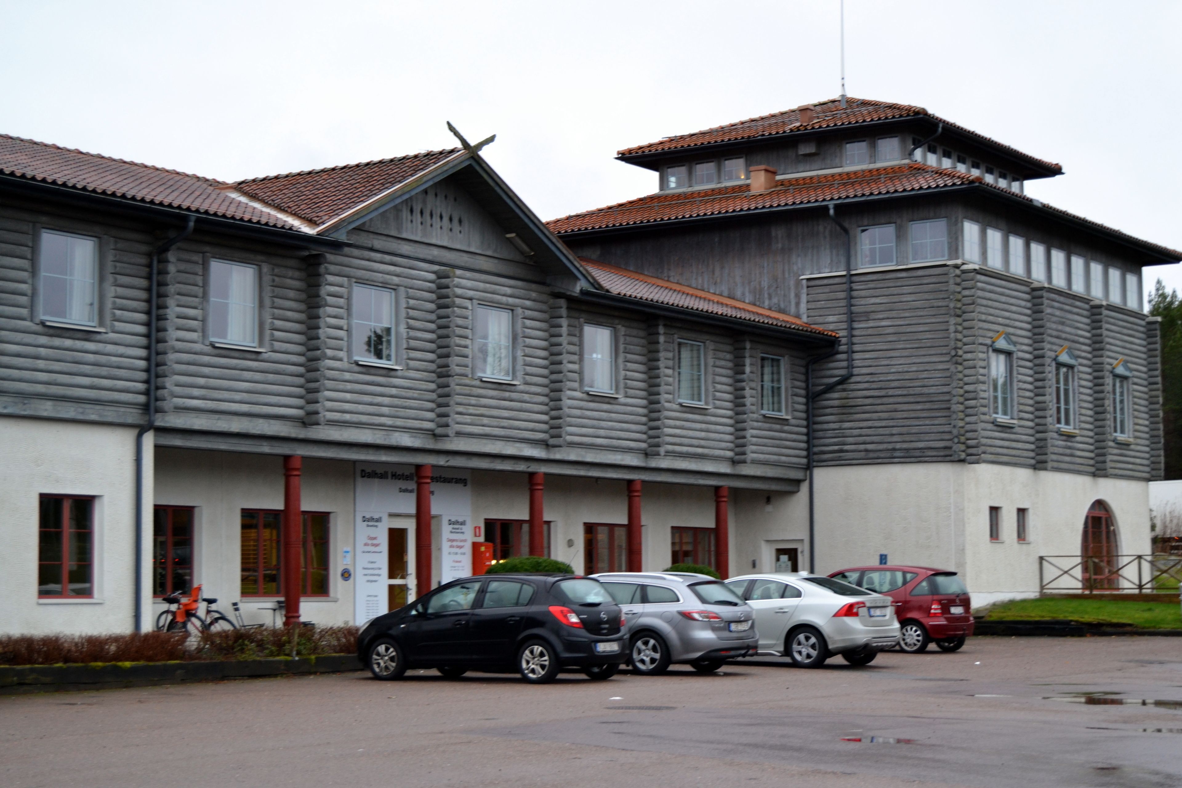 A two-story building with a traditional architectural style, featuring a mix of wooden and plaster exteriors. There are several parked cars in front of the building, and a bicycle is visible to the left.