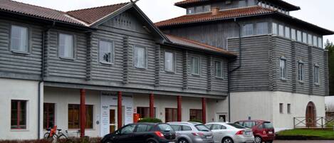 A two-story building with a traditional architectural style, featuring a mix of wooden and plaster exteriors. There are several parked cars in front of the building, and a bicycle is visible to the left.