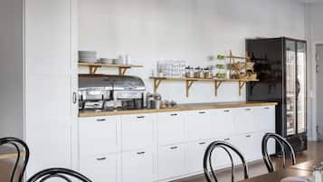 Une cuisine moderne avec des armoires blanches, un barbecue en acier inoxydable et un réfrigérateur. On y trouve des étagères avec de la verrerie et des bocaux, ainsi qu’une table en bois avec des chaises.