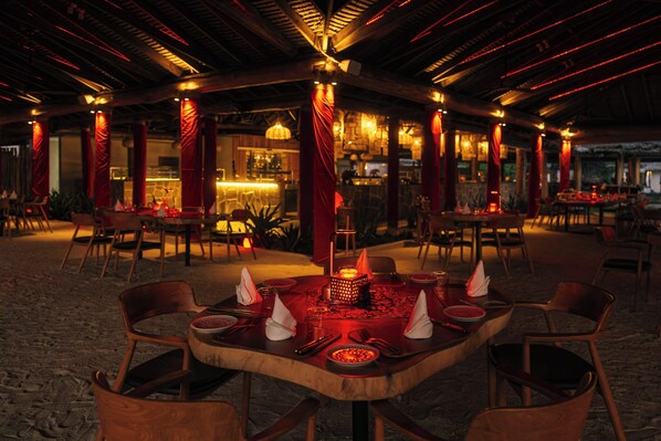 Un área de comedor al aire libre con mesas dispuestas para dos, rodeada de pilares rojos e iluminación cálida.