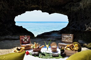 A heart-shaped opening in a rocky cliff overlooking the sea, with picnic baskets and food items on a table below. - Bawe Tropical Island (Bawe Island)