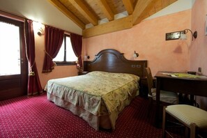 A bedroom with a wooden ceiling, a bed with a floral bedspread, a desk with a chair, and a window with curtains. - Hotel L'aiglon (Ayas)