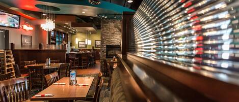 A restaurant with wooden tables and chairs, a bar area, and a wall of illuminated bottles.
