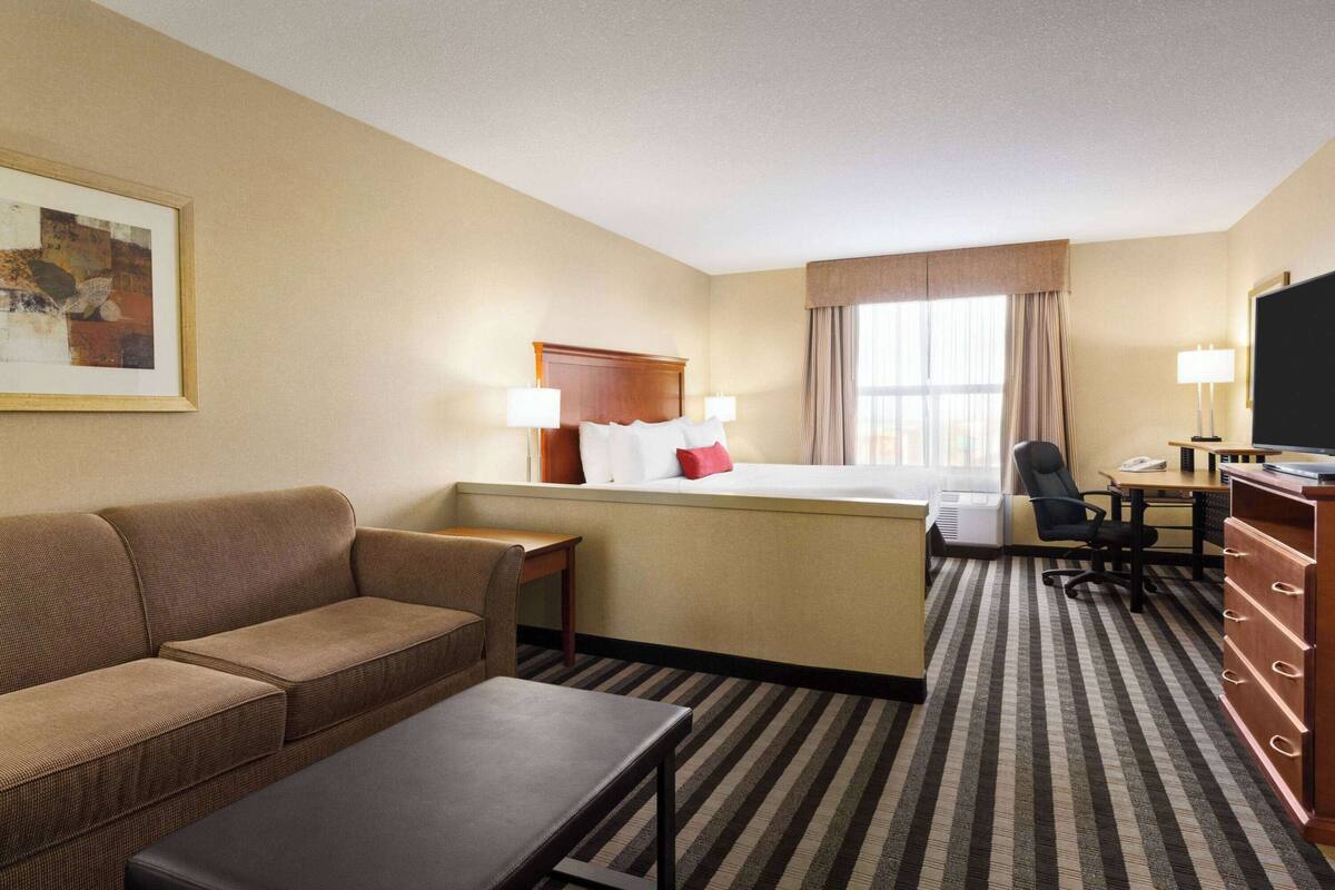 Habitación de hotel con sofá, una mesita, escritorio con silla, cama con cabecera, televisor y ventana con cortinas.
