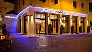 Fachada de hotel con letreros iluminados, ventanas amplias y plantas en macetas.