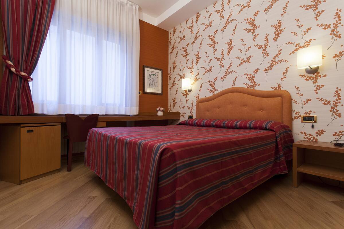 A hotel room with a bed, a desk, a chair, a nightstand, and a wall-mounted lamp.