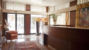 Un vestíbulo con mostrador de recepción, una silla, un jarrón grande con flores, una lámpara y un cuadro en la pared.